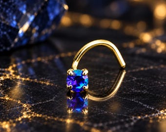 May include: A gold-toned curved nose stud featuring a single round-cut blue gemstone held in a four-prong setting. The metal post is bent into a hook shape, and the vibrant blue stone reflects light, creating a clear visual contrast against the surface.