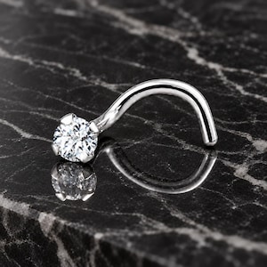 May include: A silver-toned nose screw featuring a single round, clear, faceted gemstone held in a four-prong setting. The jewelry has a curved post design and a polished metallic finish, reflecting light on its surface.