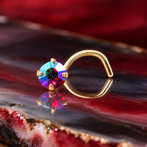 May include: A gold-toned curved nose stud featuring a round, multi-faceted crystal that displays shifting iridescent hues of red, blue, and green. The crystal is held in place by a four-prong setting, and the metal post is curved for placement.