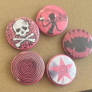 May include: A collection of five round buttons with pink and black designs. One button features a skull and crossbones, another a spiral, and a third a star. The remaining buttons have a music-themed design and a jagged pattern.