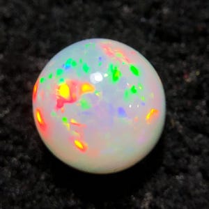 Könnte beinhalten: Ein runder, weißer Opal-Edelstein mit lebendigen Farbblitzen, darunter Rot, Grün und Gelb. Der Opal ist vor einem dunklen, strukturierten Hintergrund platziert, der seine irisierenden Eigenschaften hervorhebt.