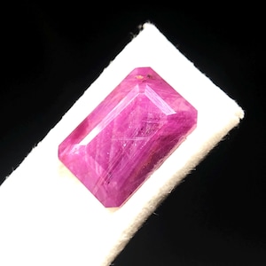 May include: A rectangular, faceted gemstone with a vibrant magenta-pink hue. The stone features a clear, polished surface with visible internal inclusions and sharp, geometric edges, showcasing a classic emerald-style cut with multiple facets.