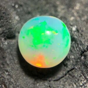 Könnte beinhalten: Ein runder Opal-Edelstein mit einem lebendigen Farbspiel. Der Opal zeigt eine Mischung aus grünen, blauen und orangen Farbtönen mit einer milchig-weißen Basis. Der Edelstein ist vor einem dunklen, strukturierten Hintergrund platziert.