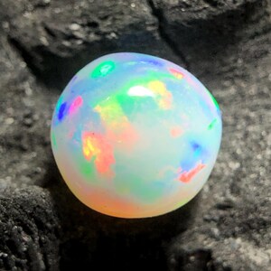 May include: A round, iridescent opal gemstone. The opal displays a milky white base color with flashes of green, blue, pink, and yellow. The gemstone is set against a dark, textured background.