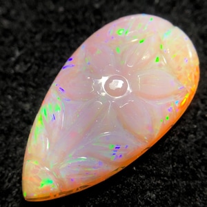 May include: A pear-shaped, carved opal gemstone with a floral design. The opal displays a play of color with hues of green, blue, and orange. The gemstone is set against a dark background.