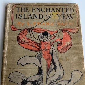 May include: A vintage hardcover book titled The Enchanted Island of Yew by L. Frank Baum. The tan cloth cover features a black and orange illustration of a figure draped in flowing fabric, with black text printed at the top of the front cover.