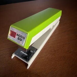 May include: A vintage ACCO 50 stapler with a lime green top and a white base. The stapler has a silver metal staple holder and the ACCO 50 logo on the side. The stapler is on a brown wooden surface.