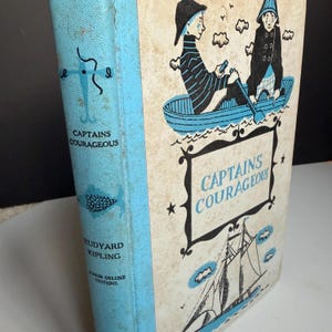 May include: A light blue and cream hardcover book titled Captains Courageous by Rudyard Kipling, Junior Deluxe Editions. The cover features a black and blue illustration of two people in a rowboat and a sailing ship, with a decorative frame.