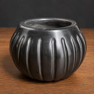 May include: A small, round, black bowl with a smooth, matte finish and a series of vertical, recessed grooves running along its exterior. The bowl features a wide, open top with a thick, rounded rim and a deep, hollow interior cavity.