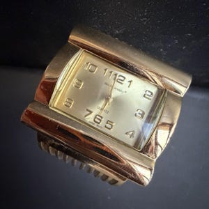 May include: A gold-toned quartz watch with a rectangular face. The watch face displays the numbers 1 through 12 in a classic font, with the brand name "Sarah Coventry" printed above the center. The watch band is integrated into the design.