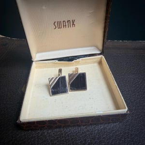 May include: Vintage cufflinks in a brown box with the brand name "SWANK" printed on the inside lid. The cufflinks are square with a black center and silver and gold accents. The box has a textured, faux-leather exterior.