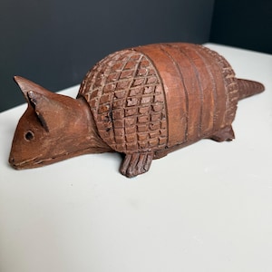 May include: A brown carved wooden armadillo figurine featuring a textured, segmented shell pattern on its back, pointed ears, and a long tail. The sculpture displays visible wood grain and hand-carved details across its entire surface.