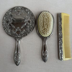 May include: A vintage vanity set featuring a round hand mirror, a hairbrush, and a comb. The set has ornate silver-toned metal frames with floral detailing. The brush has a light yellow base and bristles, and the comb is light yellow.