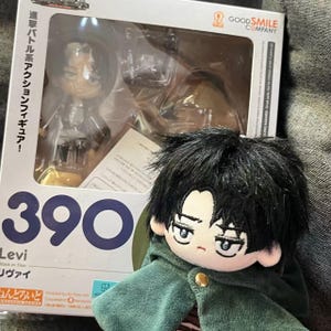 May include: A boxed Levi Nendoroid figure from the anime Attack on Titan, with the number 390 visible. Beside it is a plush doll of Levi wearing a green cloak. The box has Japanese text and the Good Smile Company logo.