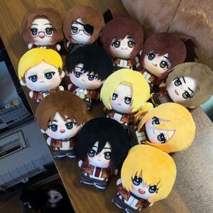 May include: A collection of plush dolls with anime-style features. The dolls have various hair colors, including brown, black, and yellow, and are dressed in brown and white outfits. Some dolls have glasses or eye patches.