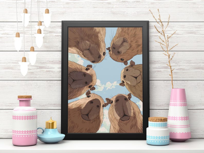 May include: A framed art print featuring a group of capybaras looking down at the viewer. The artwork has a blue sky background with fluffy white clouds. The frame is black. Decorative items include a pink and white vase, a blue candle holder, and a small jar.