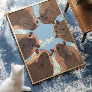 May include: A framed print featuring a group of capybaras looking down at the viewer. The capybaras are brown with light-colored faces, set against a blue sky with clouds. A white cat sits in the foreground.