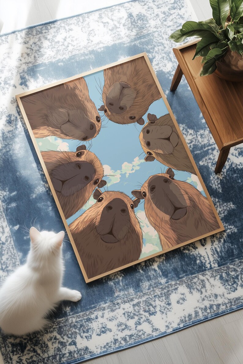 May include: A framed print featuring a group of capybaras looking down at the viewer. The capybaras are brown with light-colored faces, set against a blue sky with clouds. A white cat sits in the foreground.