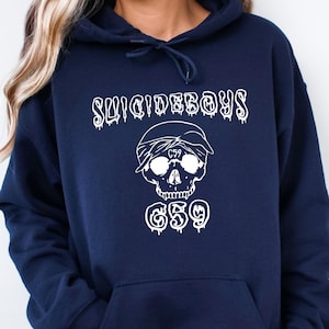 May include: Navy blue hooded sweatshirt featuring the text "SUICIDEBOYS" above a skull graphic wearing a bandana and sunglasses. Below the skull is the text "$59". The sweatshirt has a front pocket and a drawstring hood.