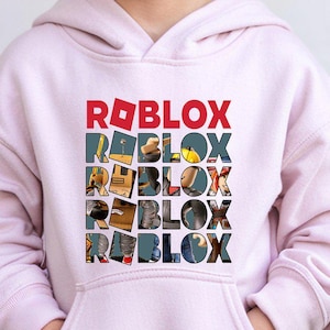 Może przedstawiać: Jasnor&oacute;żowa bluza z kapturem z napisem "ROBLOX" wydrukowanym pionowo w kolorowym wzorze. G&oacute;rny napis "ROBLOX" jest czerwony, pozostałe to kolaż obraz&oacute;w. Bluza ma przednią kieszeń.