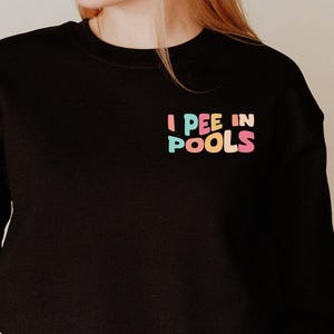 May include: Black sweatshirt with the text "I PEE IN POOLS" in pastel colors on the upper left chest. The sweatshirt is long-sleeved and has a crew neck. The model is wearing blue jeans.
