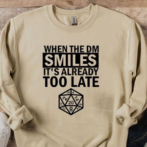 May include: A tan sweatshirt with the text "WHEN THE DM SMILES IT'S ALREADY TOO LATE" in black, along with a D20 die graphic. The sweatshirt is paired with blue jeans and white sneakers on a wooden surface.