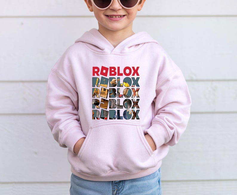 Może przedstawiać: Jasnor&oacute;żowa bluza z kapturem z napisem "ROBLOX" wydrukowanym pionowo w kolorowym wzorze. G&oacute;rny napis "ROBLOX" jest czerwony, pozostałe to kolaż obraz&oacute;w. Bluza ma przednią kieszeń.