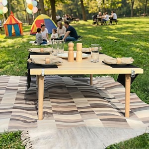 May include: A light-colored wooden picnic table with folding legs, set on a striped blanket. The table is set with plates, napkins, glasses, and salt and pepper shakers. A family is picnicking in the background.