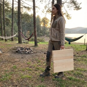 May include: A light-colored, wooden folding table with a white handle is being carried outdoors. The table is made of several wooden planks. The person is wearing a tan jacket and pants, and hiking boots. The background includes trees, hammocks, and a lake.