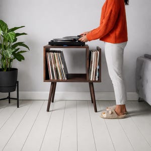 May include: A dark brown wooden record player stand with angled legs. The stand has two compartments filled with vinyl records. A black record player sits on top. A person wearing an orange jumper and white trousers is standing next to the stand.
