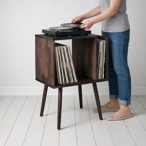 May include: A dark brown wooden record player stand with a built-in storage compartment for vinyl records. The stand has a mid-century modern design with angled legs. A person is placing a record on the turntable.