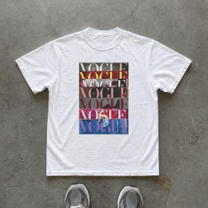 May include: White t-shirt featuring a rectangular graphic with the word "VOGUE" stacked multiple times in various colors. The shirt has short sleeves and a crew neck. The image is taken from a top-down perspective.