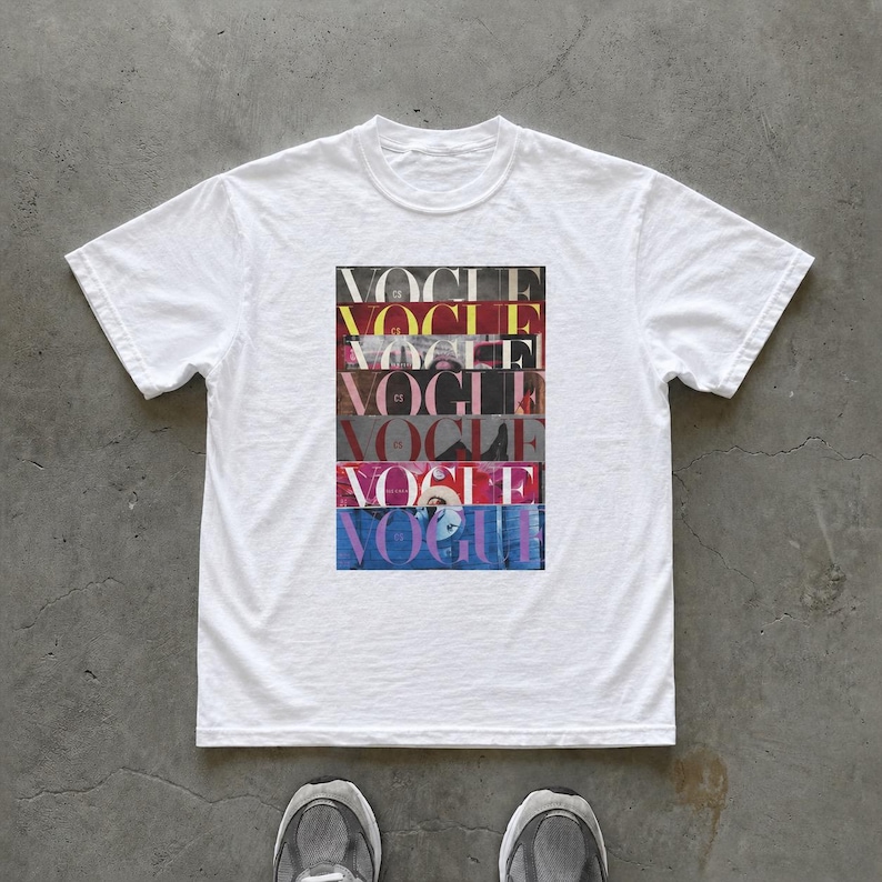May include: White t-shirt featuring a rectangular graphic with the word "VOGUE" stacked multiple times in various colors. The shirt has short sleeves and a crew neck. The image is taken from a top-down perspective.