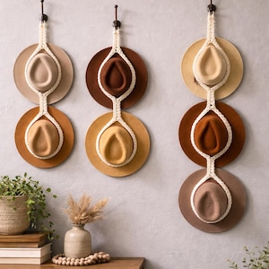 May include: Three hat racks, each holding two hats, are mounted on a light gray wall. The hats are in shades of brown and beige, suspended by cream-colored macrame. A wooden table with decor is in the foreground.