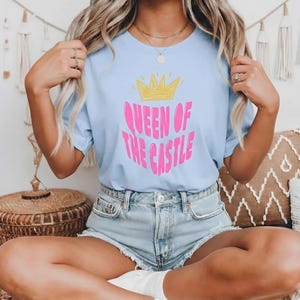 Può includere: T-shirt azzurra con la scritta rosa "QUEEN OF THE CASTLE" sormontata da una corona gialla. Un top casual.