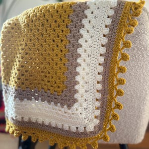 Puede incluir: Una manta de ganchillo hecha a mano con un patrón de cuadrados de la abuela. La manta presenta un diseño de bloques de color con cuadrados de color amarillo mostaza, blanco y topo. El borde es de color amarillo mostaza con un borde festoneado. La manta está sobre una silla.