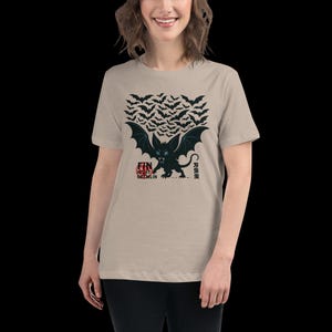 May include: Beige t-shirt featuring a black and teal bat-like creature with large wings, a menacing expression, and a swarm of bats above. The text "FIN NIGHT GREMLIN" is printed below the creature.