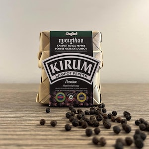 Peut inclure: Un paquet rectangulaire de poivre noir de Kampot enveloppé dans une matière en feuille de palmier tissée beige clair. L'étiquette porte le texte Confirel, Kampot Black Pepper, Poivre Noir de Kampot et Kirum Kampot Pepper. Des grains de poivre sont éparpillés.