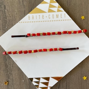 May include: Two red and gold beaded hair clips on a white background with the text "Brite Comet" printed on the surface.