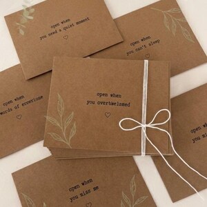 May include: A collection of brown cards tied with white string, each featuring text and a leaf design. The cards bear phrases such as "open when you need a quiet moment" and "open when you miss me". The cards are a warm brown colour.