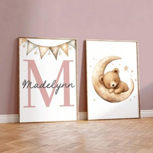 May include: Two framed nursery art prints. One features a pink letter "M" with the name "Madelynn" and a banner of stars. The other shows a sleeping teddy bear on a crescent moon with stars.
