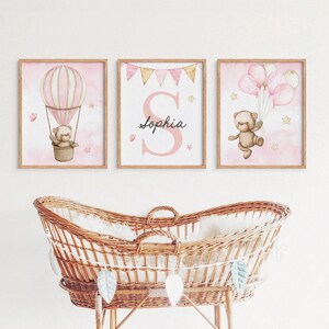 May include: Three framed nursery art prints featuring a hot air balloon, a teddy bear with balloons, and the name "Sophia". The prints have a pink and white color scheme with star and butterfly accents. The art is displayed above a wicker bassinet.