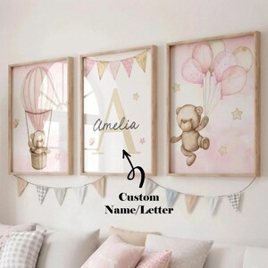 May include: Three framed nursery art prints with a soft pink and beige color scheme. One print features a teddy bear in a hot air balloon, another has a large letter "A" and the name "Amelia", and the third shows a teddy bear with balloons. A banner hangs below.