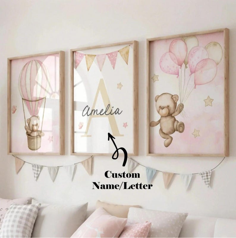 May include: Three framed nursery art prints with a soft pink and beige color scheme. One print features a teddy bear in a hot air balloon, another has a large letter "A" and the name "Amelia", and the third shows a teddy bear with balloons. A banner hangs below.