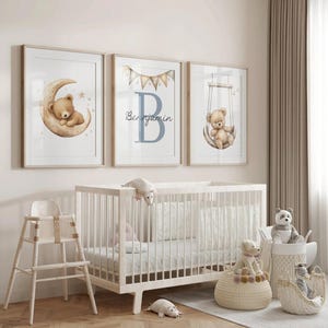May include: Three framed nursery art prints featuring watercolor teddy bears. One shows a bear on a crescent moon, another has a large blue letter "B" with the name "Benjamin", and the third shows a bear on a swing. A white crib and stuffed animals are in the foreground.