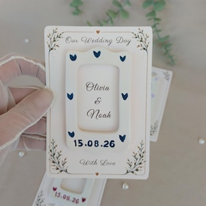 Può includere: Una partecipazione di nozze rettangolare con una cornice centrale bianca decorata con cuori blu scuro, i nomi Olivia e Noah e la data 15.08.26. La carta riporta le scritte Our Wedding Day e With Love, arricchita da illustrazioni botaniche.