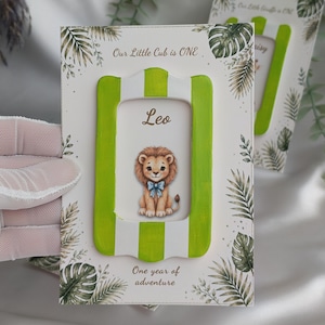 May include: A personalized photo frame with a green and white striped border. The frame features a cartoon lion with a blue bow tie and the name "Leo". Text at the top reads "Our Little Cub is ONE" and at the bottom "One year of adventure".