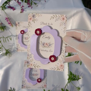 Könnte beinhalten: Blumenkarten mit einem lavendelfarbenen, gewellten Rahmen und roten Blumenakzenten. Auf den Karten steht "Bride to be", "Emily" und "Celebrating Love". Der Hintergrund zeigt ein Blumenmuster mit rosa und lila Rosen.