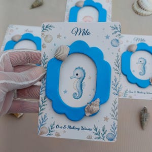 Könnte beinhalten: Eine Sammlung personalisierter Fotorahmen mit maritimem Thema. Jeder Rahmen hat einen blauen, gewellten Rand, eine Seepferdchen-Illustration und den Namen "Milo" oder "Aria". Muscheln und Wassermotive verzieren die Rahmen.
