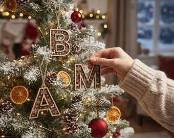 May include: Three wooden letter ornaments shaped as A, B, and M, featuring intricate laser-cut snowflake and deer patterns. Each letter is suspended by a thin twine string. The ornaments are displayed on a flocked Christmas tree with pinecones.